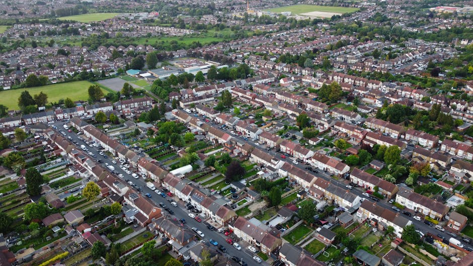An aerial view of a residential neighbourhood showing a row of terraced houses with front gardens, paved streets, and numerous parked cars lining both sides. The image includes greenery such as trees and lawns, with some houses featuring visible packaging materials like cardboard boxes and wrapped furniture in driveways or on the pavement, indicating a home relocation or packing process. The scene is well-lit with natural daylight, capturing the organized layout of houses and streets in a typical suburban area, supporting the logistics of furniture transport and house removals. This visual exemplifies a typical setting where Brent Man and Van facilitates house removals, highlighting the importance of careful packing, loading, and transport within London's suburban streets, particularly in the HA9 area for smooth moving and relocation services.