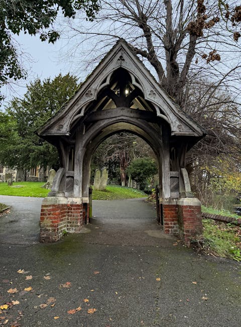 A wooden Gothic-style archway structure with intricate carved details and a pointed gabled roof, situated at the entrance of a park or garden. The archway is supported by brick pillars at its base, with a paved pathway leading through it into a green area with grass, trees, and headstones visible in the background. Surrounding the scene, some trees have no leaves while others have green foliage, indicating late autumn or early winter. The sky is overcast, casting diffuse natural light over the scene. This structure may serve as an entrance or a decorative feature in a historic park, and it is part of an outdoor setting where house removals or relocation services might operate during property moves involving nearby residential or historic locations. Brent Man and Van provides professional removals services in areas including Brents' streets, which might involve transporting furniture or household items past such landmarks.