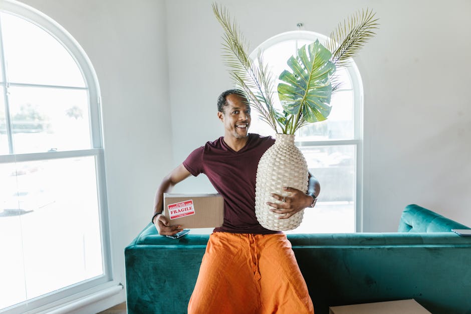 A man with dark skin, wearing a purple t-shirt and orange trousers, stands indoors in front of a large window. He is smiling and holding a large white textured ceramic vase filled with green tropical 