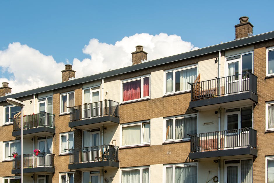 A close-up view of the exterior facade of a high-rise residential building with multiple floors, each featuring balcony spaces enclosed with black metal railings and glass sliding doors. Some balconie
