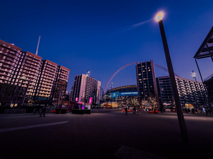 A wide-angle view of an outdoor city scene at dusk, featuring modern high-rise buildings with illuminated windows and a large Ferris wheel in the distance. In the foreground, a paved open area has lampposts casting bright light, with a few pedestrians walking across. To the right, part of a building's exterior with a metal canopy is visible. The scene includes lifted furniture and boxes wrapped in plastic and cardboard, being loaded into a van by movers, with equipment such as trolleys and packing blankets indicating a home relocation or furniture transport process. Brent Man and Van's logistics are implied through the context of house removals and moving services, subtly incorporated into the urban setting. The overall atmosphere suggests the preparation or active phase of packing and loading during an evening move. The environment is well-lit with city lights, emphasizing the busy, organized nature of a professional removal service operating in an urban residential or commercial area.